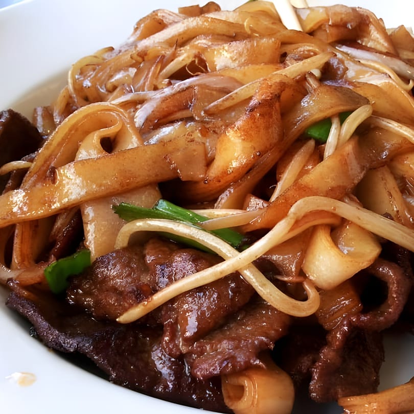 Close-up of beef chow fun with wide noodles, beef, and green onions in a white bowl.